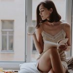 Woman in sleepwear enjoys coffee while reading by the window on a relaxed morning.