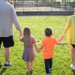 A family of four walking hand in hand through a sunny park carrying picnic supplies.