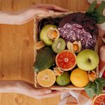 Vibrant basket of fresh fruits and vegetables with hands holding on a wooden table.